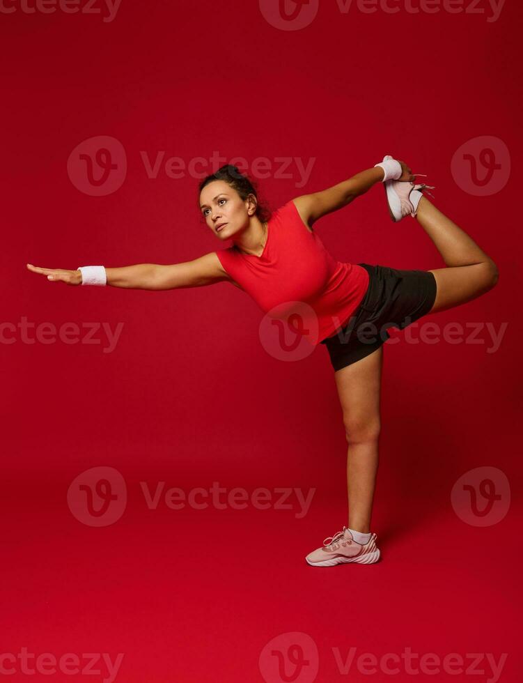 Full body size portrait of an active dark-haired Hispanic athletic woman stretching her body whiles exercising against red background with copy space photo