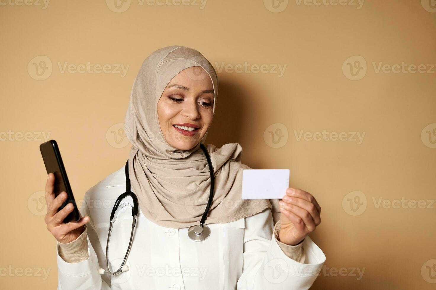 Attractive Arab doctor with covered head in hijab holding a cell phone and a blank plastic card, standing on beige background with copy space. Insurance and healthcare concept photo