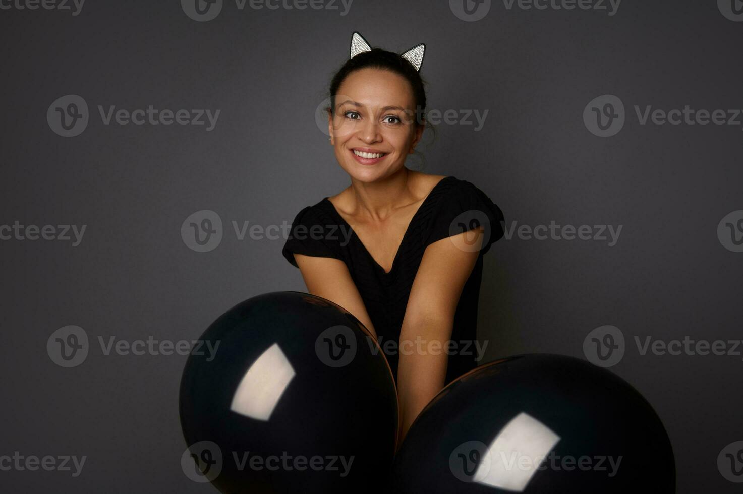 Latin American beautiful woman dressed in black, wearing hoop with cat ears, smiles toothy smile holds black air balls in front of her, looks at camera against gray background. Halloween, Black Friday photo