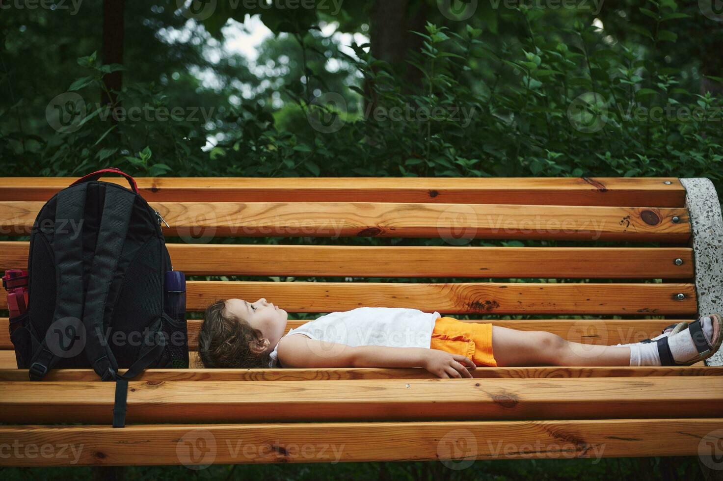 Side view of Caucasian lovely baby girl, 4 years old, adorable little kid girl lying on a wooden bench in the city park photo