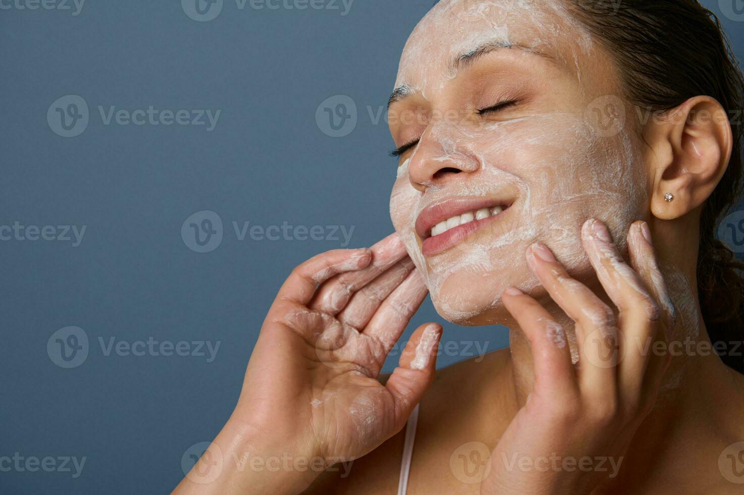 Beautiful delighted woman with perfectly healthy skin, holding hands on her face, performing smoothing massage, removing makeup and cleansing face with exfoliating scrub and facial foam cleanser. photo