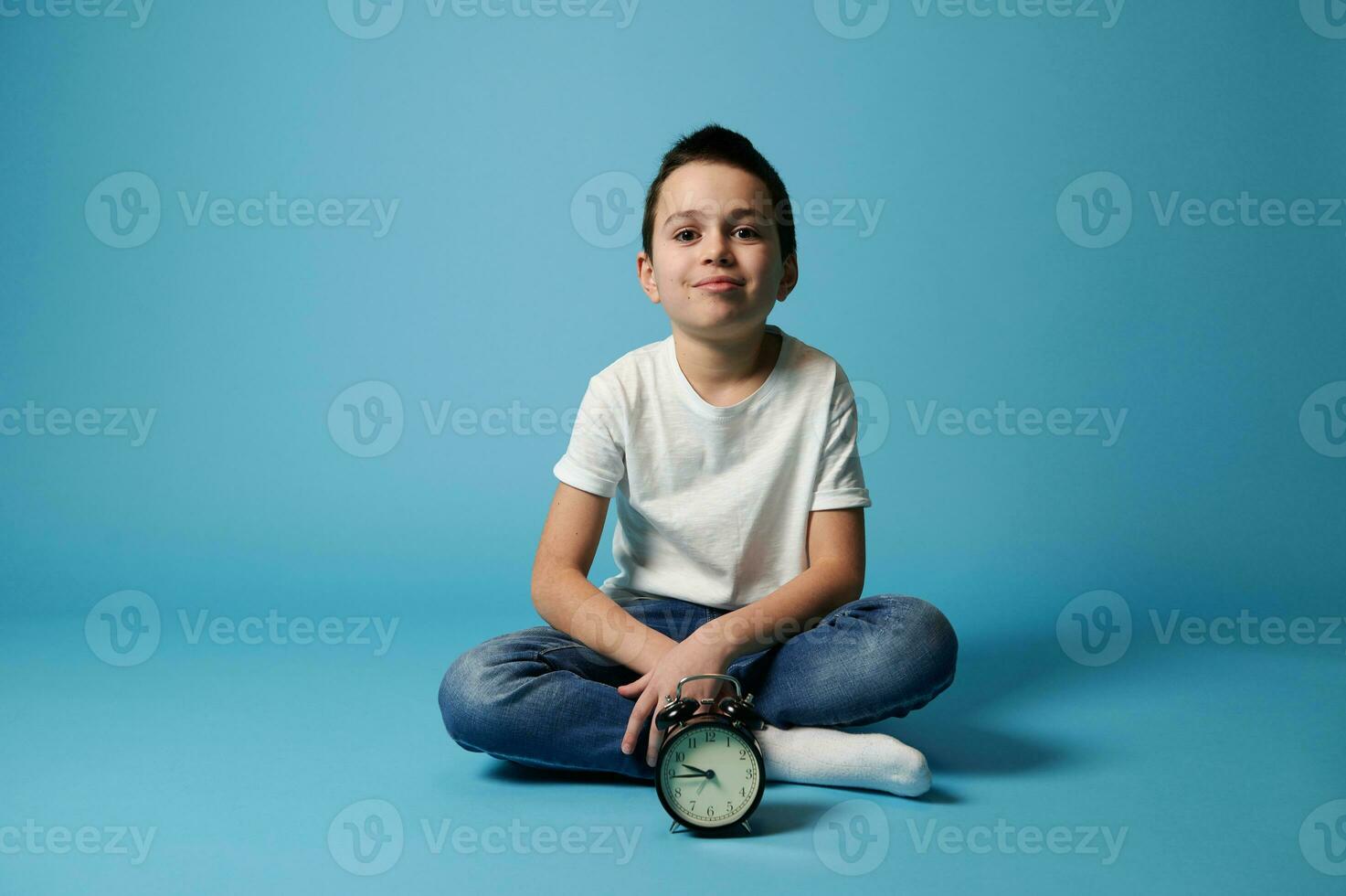 Nice boy in white t-shirt and jeans sitting on a blue background with copy space behind an alarm clock. photo