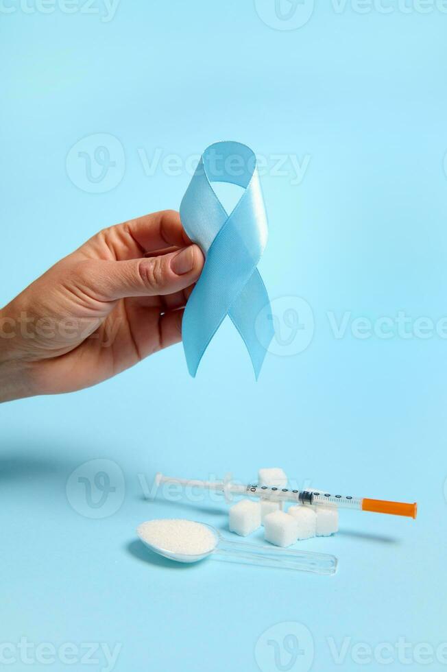 Close-up of hand holding blue ribbon, symbol of World Diabetes Awareness Day on blue background with insulin syringe and pure refined white sugar cubes. Concept of dangers of eating white sugar photo