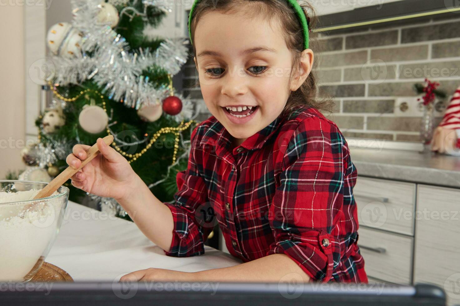 Close-up of a little vlogger, European beautiful baby girl mixing flour in glass bowl, preparing dough, talking by video call, recording video blog against a Christmas tree background at home kitchen photo