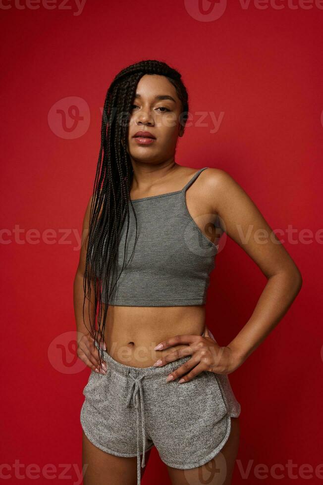 Self-confident beautiful young African American woman in gray sportswear posing with arms on waist against a red colored background with copy space for ad photo