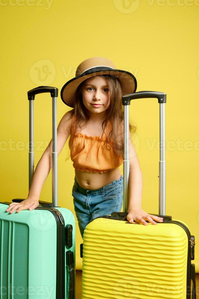 Fashionable kid girl in straw hat, with vibrant light green and yellow suitcases, travelling abroad, isolated background photo