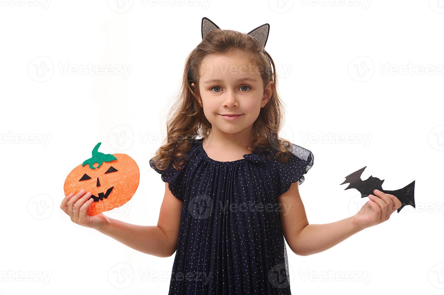 bonito pequeño niña con un aro en el formar de gato orejas, vestido en un hermosa noche vestido, sostiene corte de fieltro calabaza y hecho a mano murciélago, sonrisas mirando a cámara. aislado en blanco antecedentes Copiar espacio foto