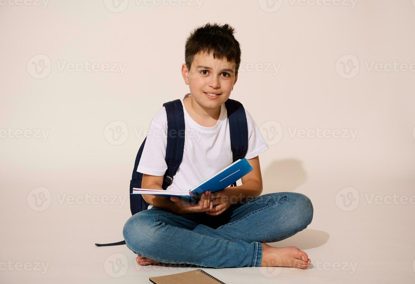 Hispanic teen boy, smart student smiles broadly at camera, holding copybook, sitting in lotus pose over white background photo