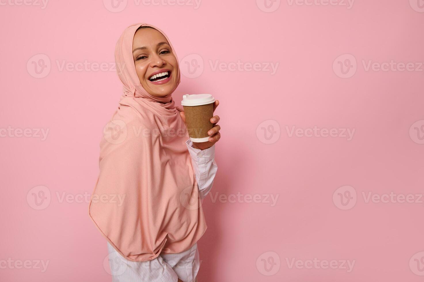Arabic Muslim woman with covered head in pink hijab holds disposable cardboard takeaway cup, smiles toothy smile, looking at camera, standing three quarters against colored background with copy space photo