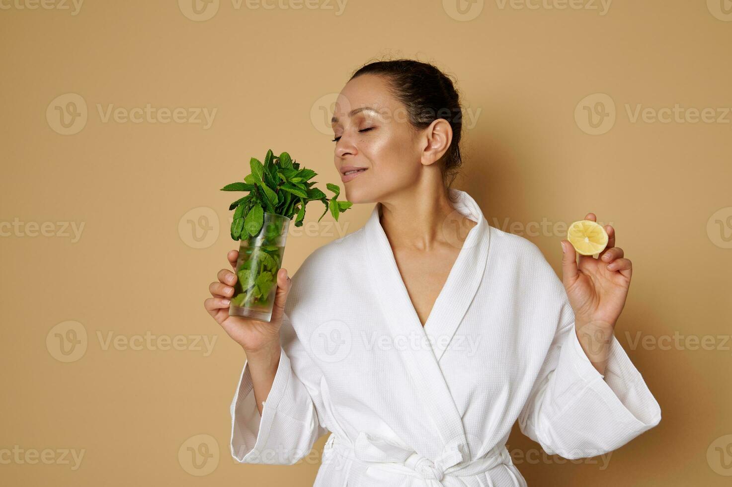 Isolated portrait on beige background of a young beautiful woman with clean skin holding a half lemon in one hand and enjoying the scent of mint in the other hand. photo