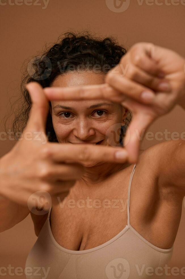 Close-up of a cheerful dark-haired curly woman wearing underclothing, smiles looking at camera through a finger frame, isolated over beige background with copy space for ad photo