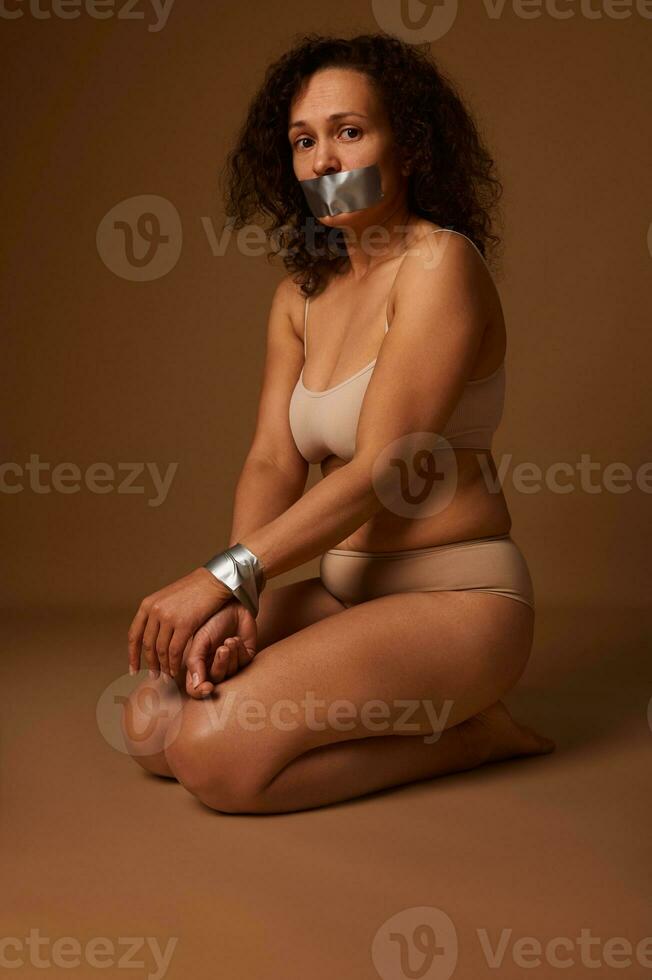 Social portrait of full-length scared woman with her hands tied, looking desperately with hope for help in eradicating aggression and violence against women. Isolated on beige background, copy space photo