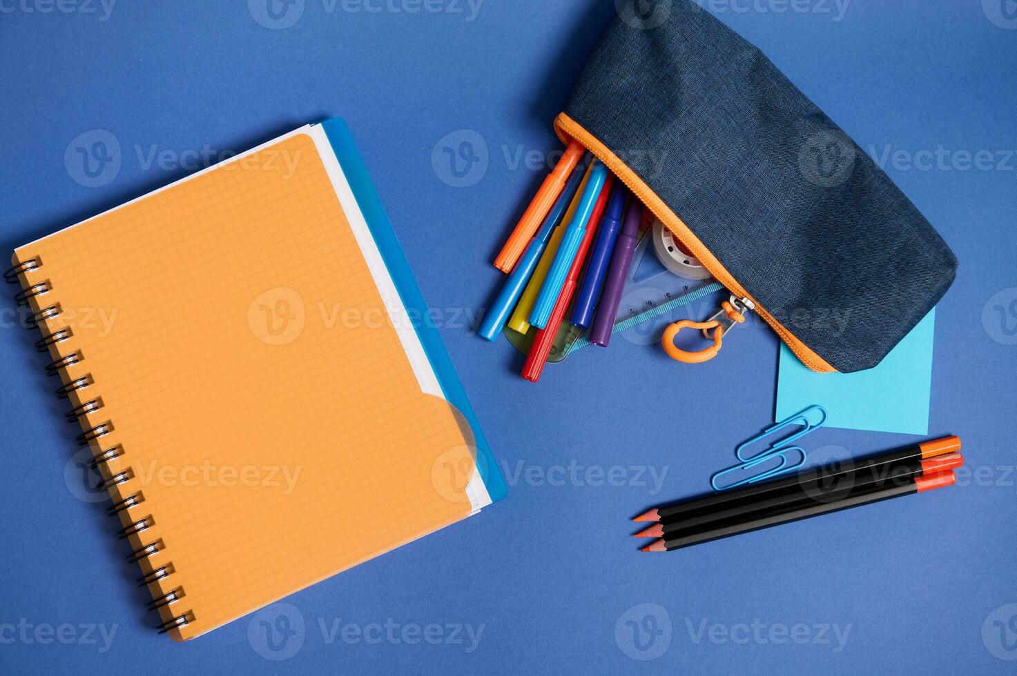 Flat lay with a copybook, organizer, colorful pencils and pencil case with falling out school supplies , in blue and orange contrast shadows isolated on blue background with copy space photo