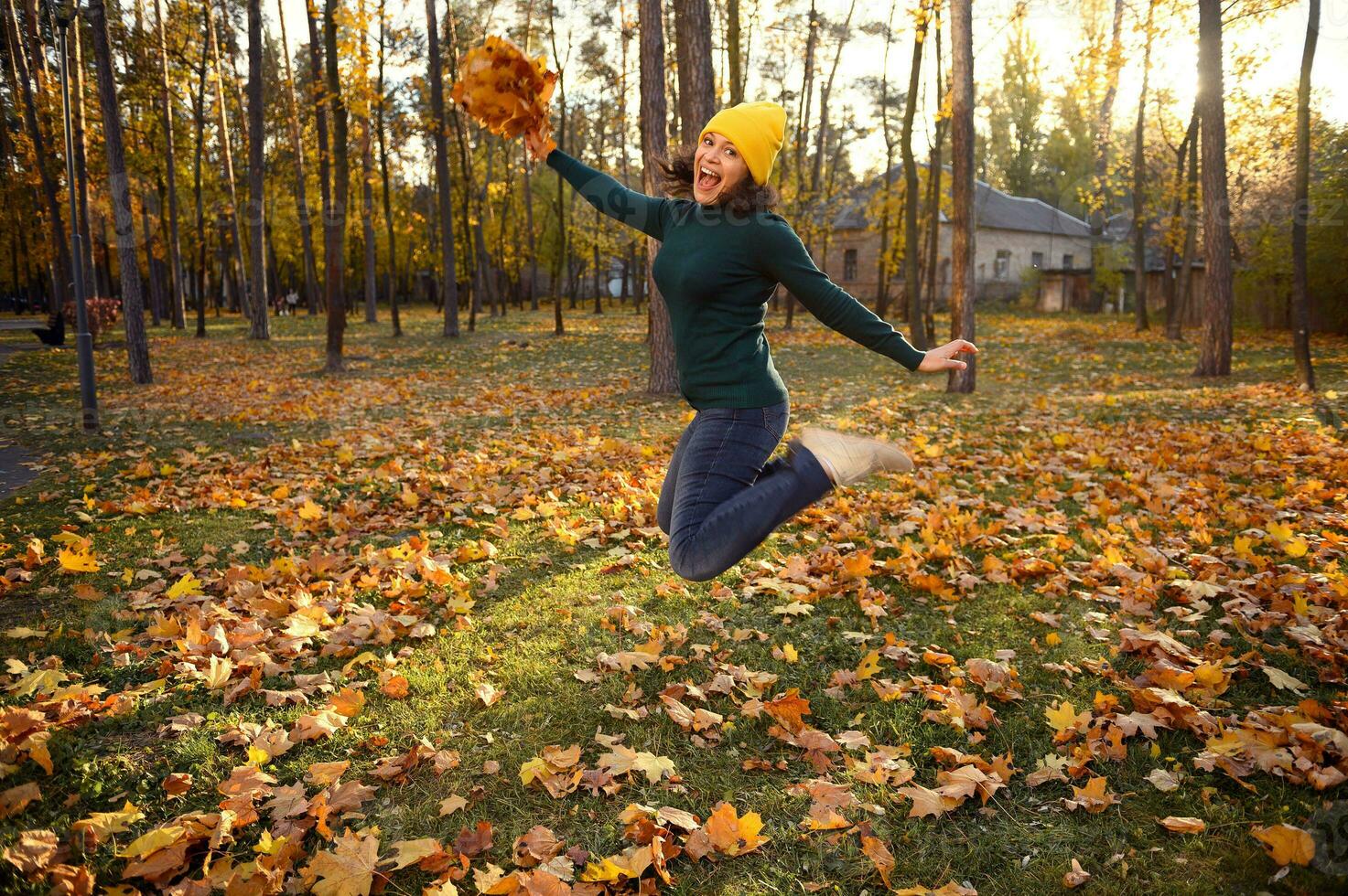 Beautiful woman in warm green pull-over, yellow woolen and casual denim hat jumping high with a bouquet of beautiful dry autumn maple leaves in hands, enjoying beautiful autumn day at sunset in park photo