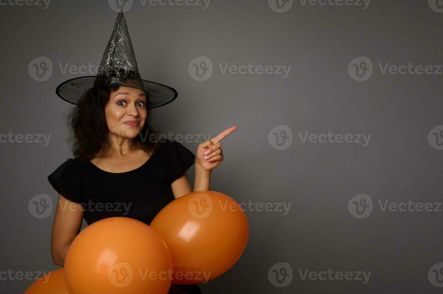 Cheerful joyful happy Hispanic woman in wizard hat, dressed in black, holds orange inflated air balloons and points on a copy space on gray background. Halloween party concept photo
