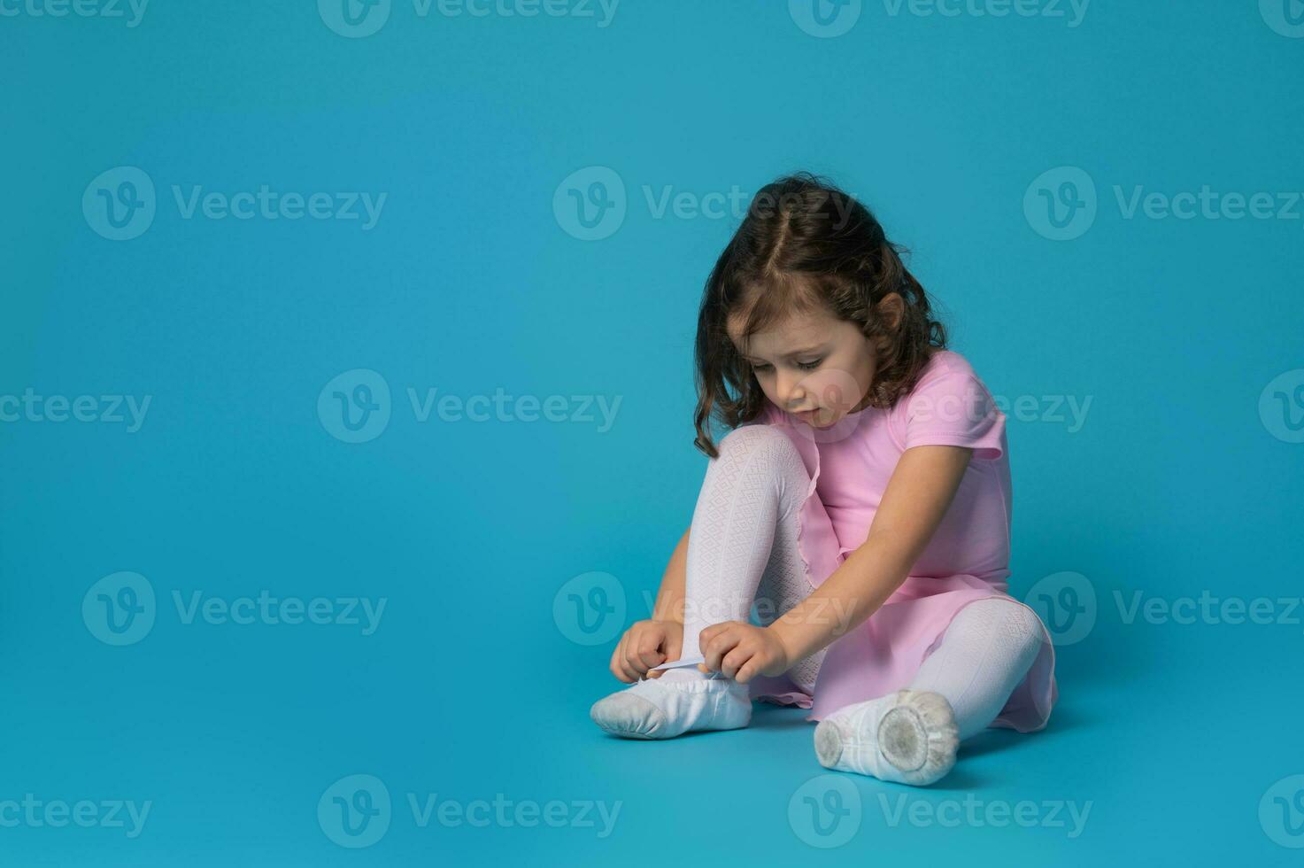 Cute ballerina focused on tying shoelaces on ballet shoes sitting on blue background photo