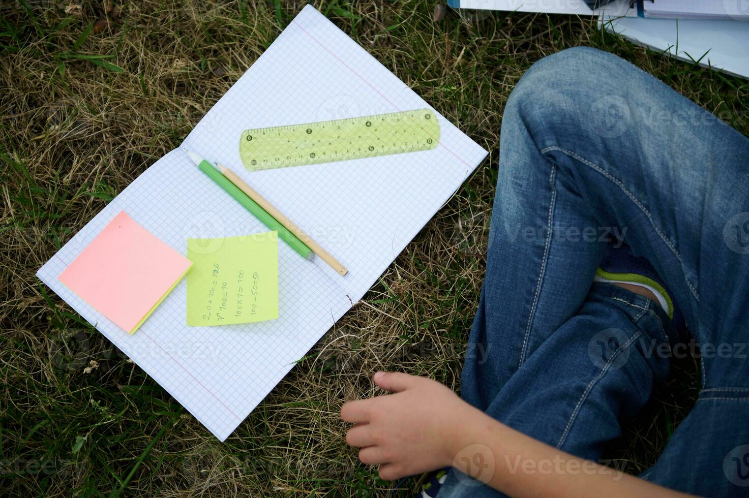 Flat lay of open notebook with colorful note papers with arithmetic problems, pencil, ruler and pen in the middle of notebook lying on green grass in park, choolboy sitting next to him doing homework photo