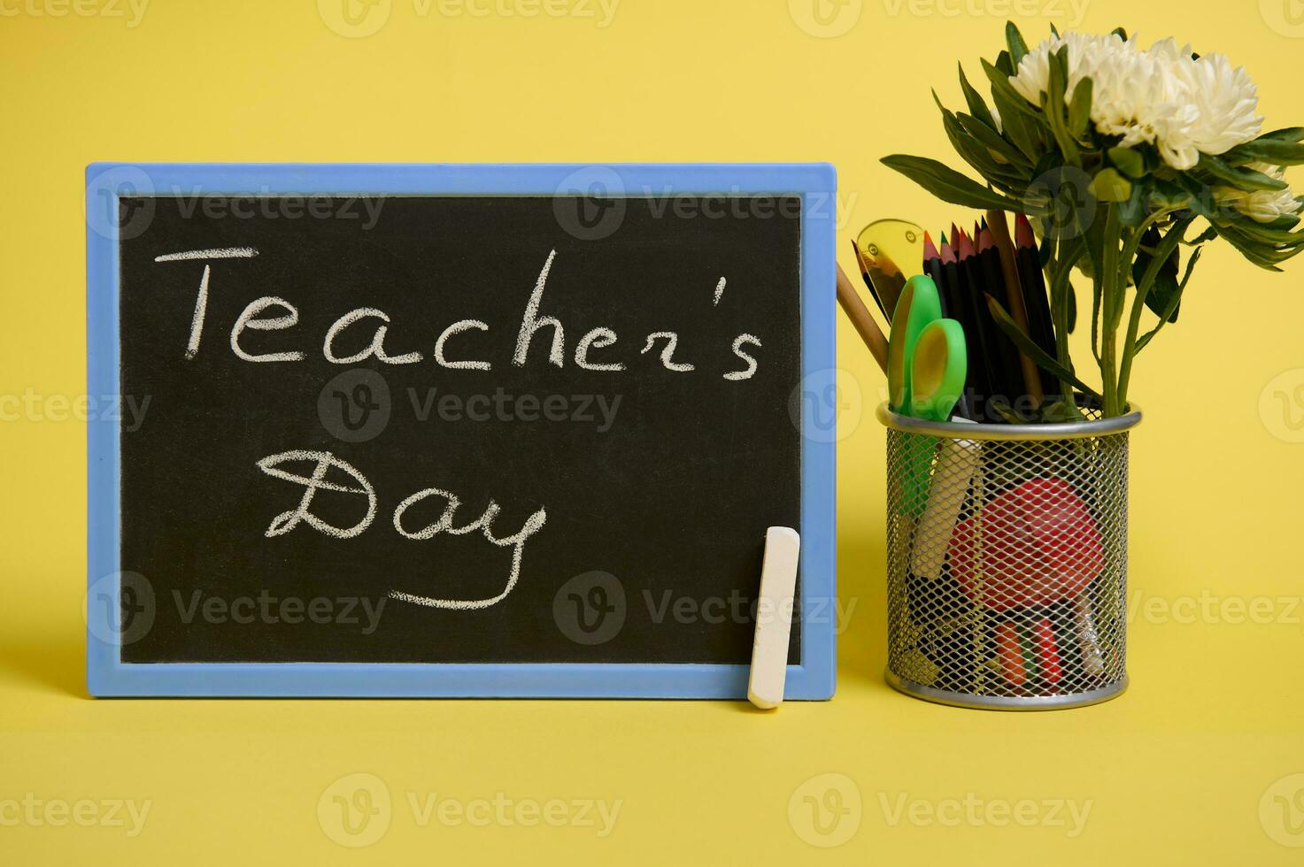 Stand for pens and pencils on the desktop on a yellow background with copy space. Holder with colored school supplies and aster flower and chalk board with lettering Teacher's Day photo