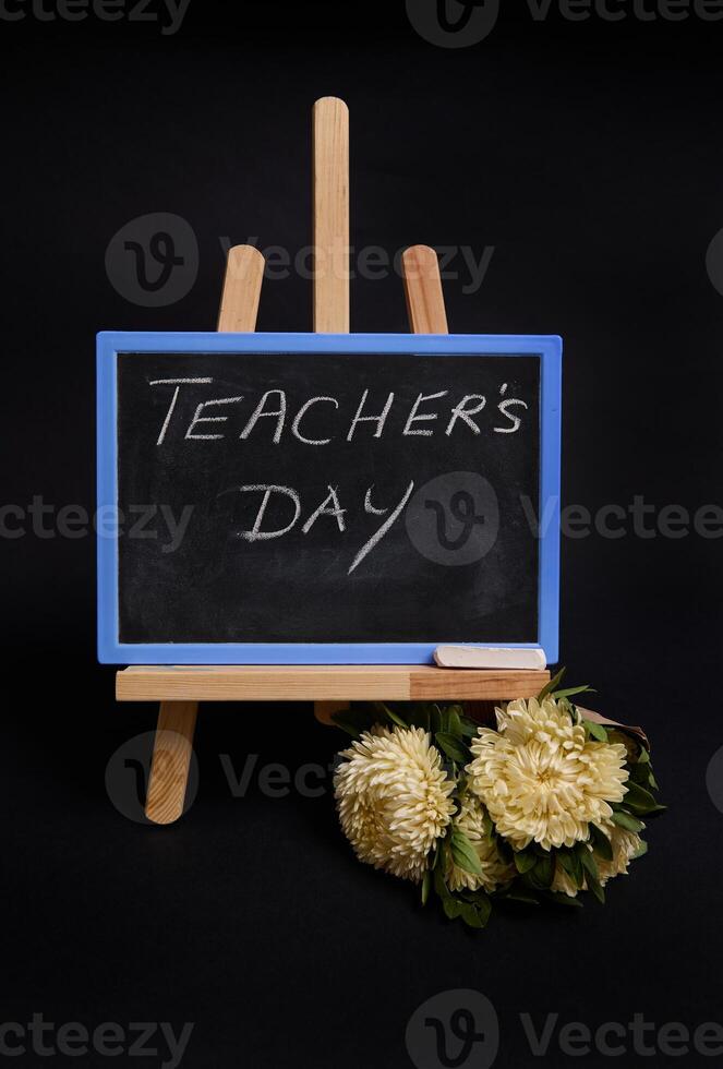 Close-up of a chalk blackboard with lettering Teacher's Day, standing on a wooden table easel, next to black alarm clock, isolated on black background with copy space. photo