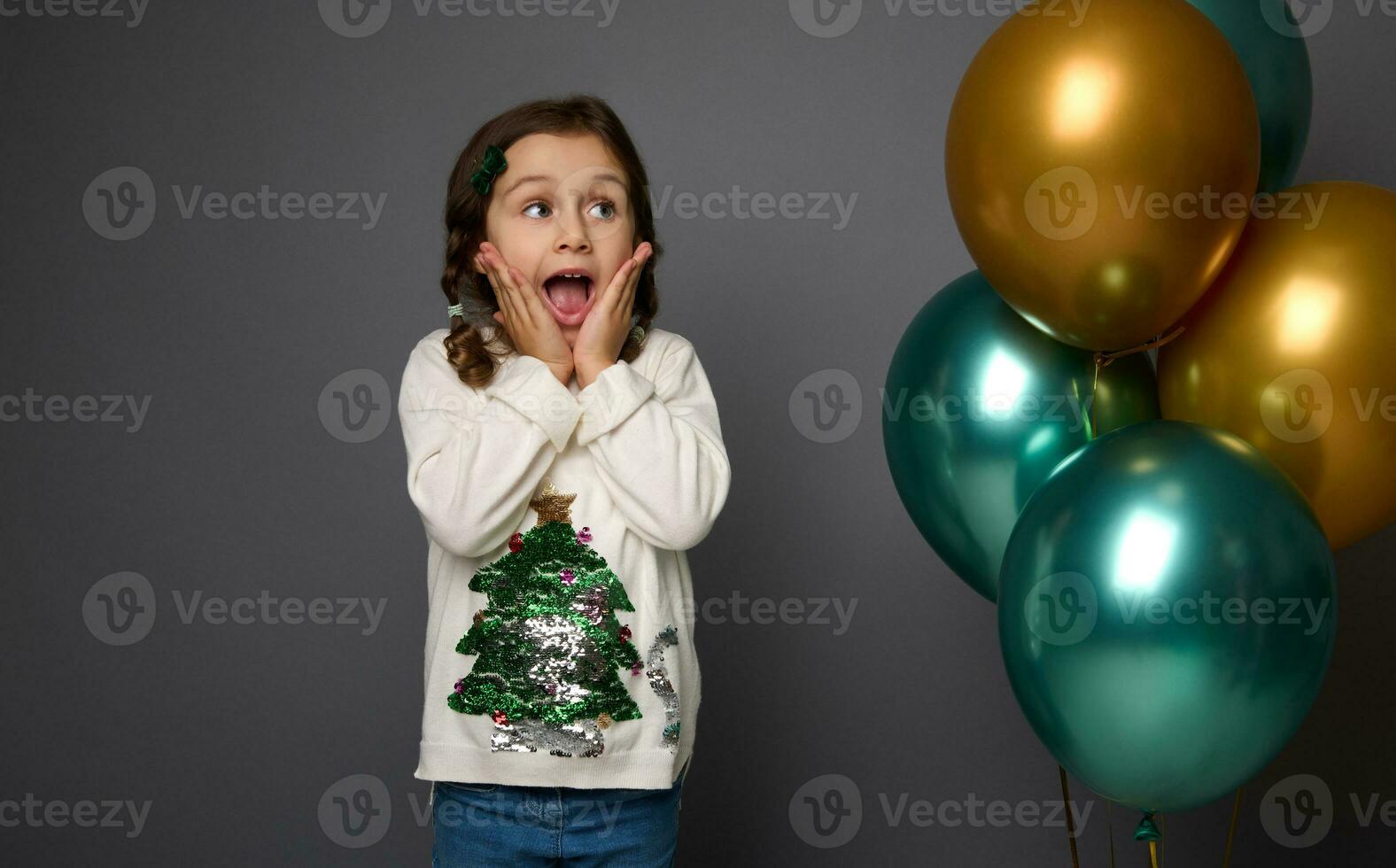 Happy surprised baby girl in sweater with shiny Christmas tree, rejoicing standing near inflated golden and green metallic air balloons against gray background with copy space. New Year concept photo