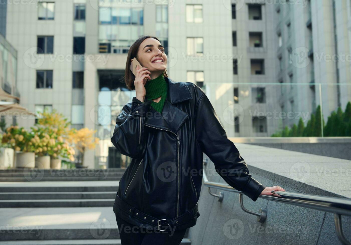 Attractive woman with stylish short hair talking on mobile phone standing outdoor against modern corporate high-rise buildings. Digital gadgets and communication concepts in contemporary life photo