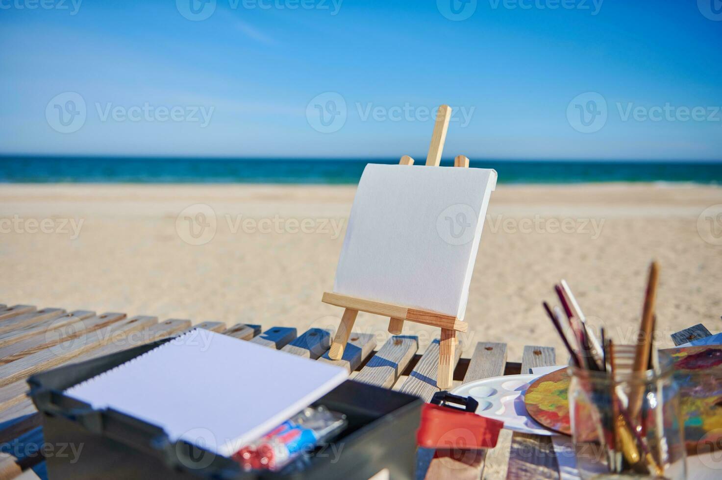 Blank white canvas on a wooden table easel next to paints and brushes on a wooden deck chair on the beach against the background of beautiful blue sea photo