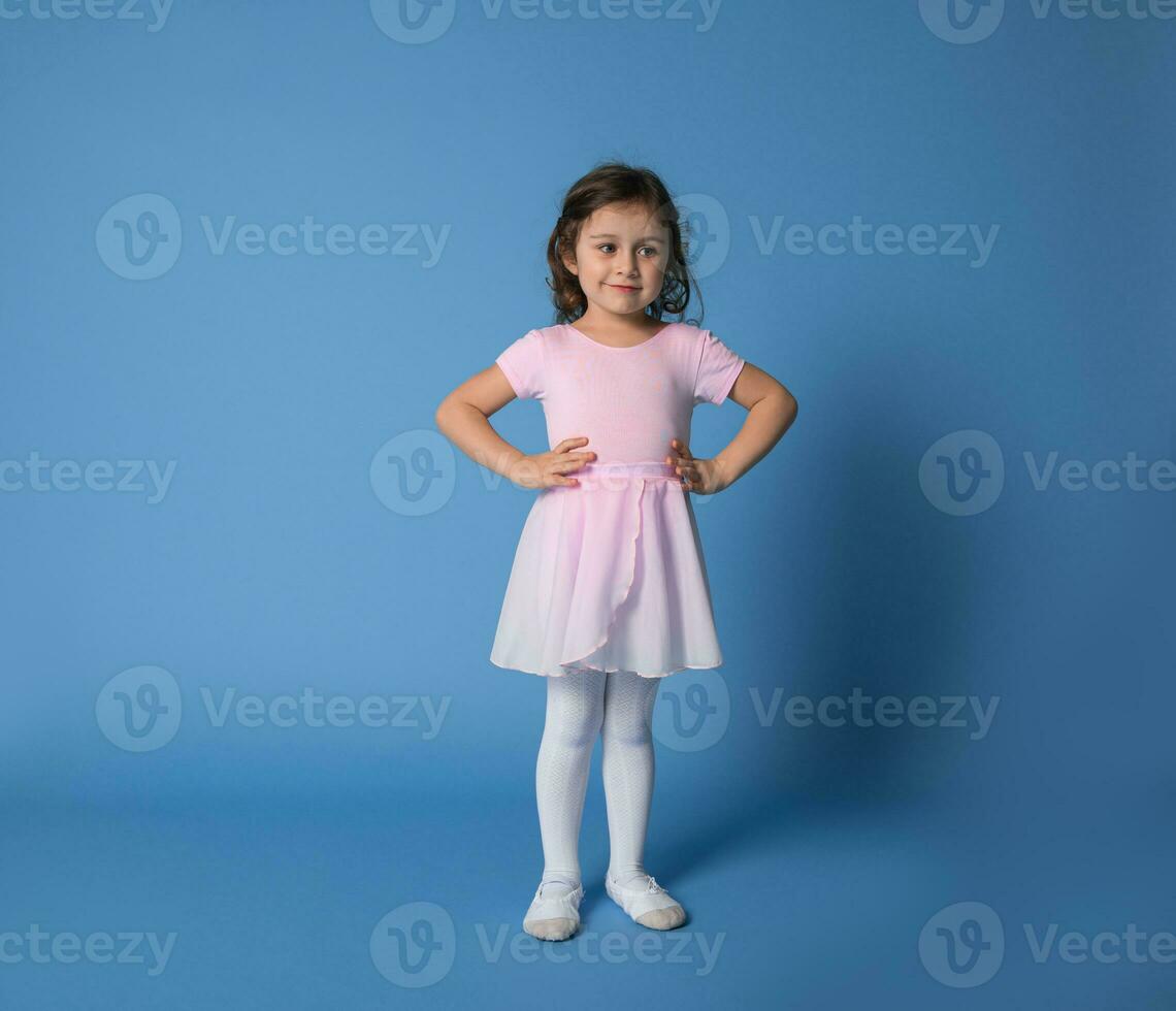 Cute girl ballerina dressed in pink uniform posing into the camera with arms on waist photo