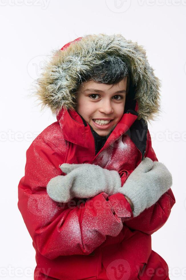 aislado retrato en blanco antecedentes de un hermoso europeo elemental años chico vistiendo un brillante rojo chaqueta con un cubierto de nieve capucha mirando a el cámara, abrazando él mismo, temblando desde el frío foto