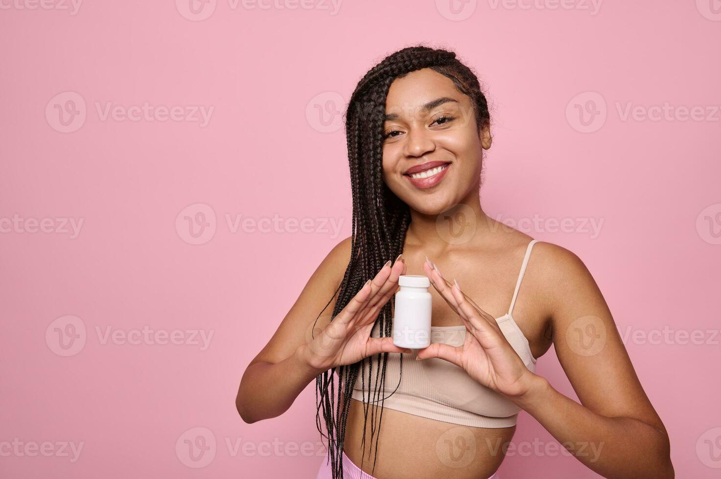 Charming African woman smiles toothy smile, holding a bottle of dietary supplements or vitamins for the beauty and health of skin and hair, posing on a pink background with space for advertising text. photo