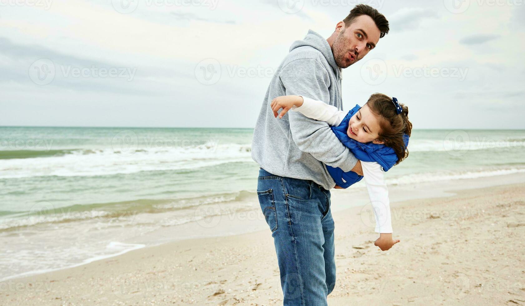 hermoso padre participación su pequeño linda hija en su brazos y jugando juntos en el playa en un hermosa frio temprano verano día. del padre día concepto foto