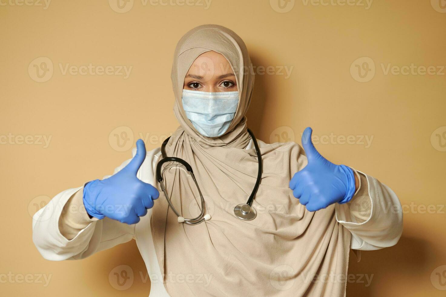 Young medic wearing hijab and medical mask showing thumbs up gesturing with hands in medical protective gloves photo