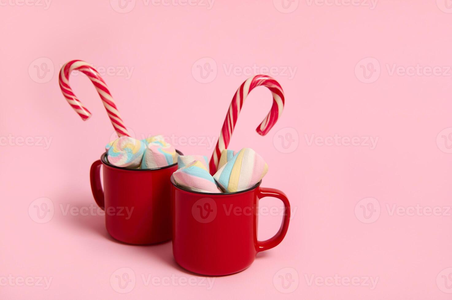 Marshmallows and candy cane in cup of hot chocolate in red mugs isolated over pink pastel background with copy space for Christmas ad photo