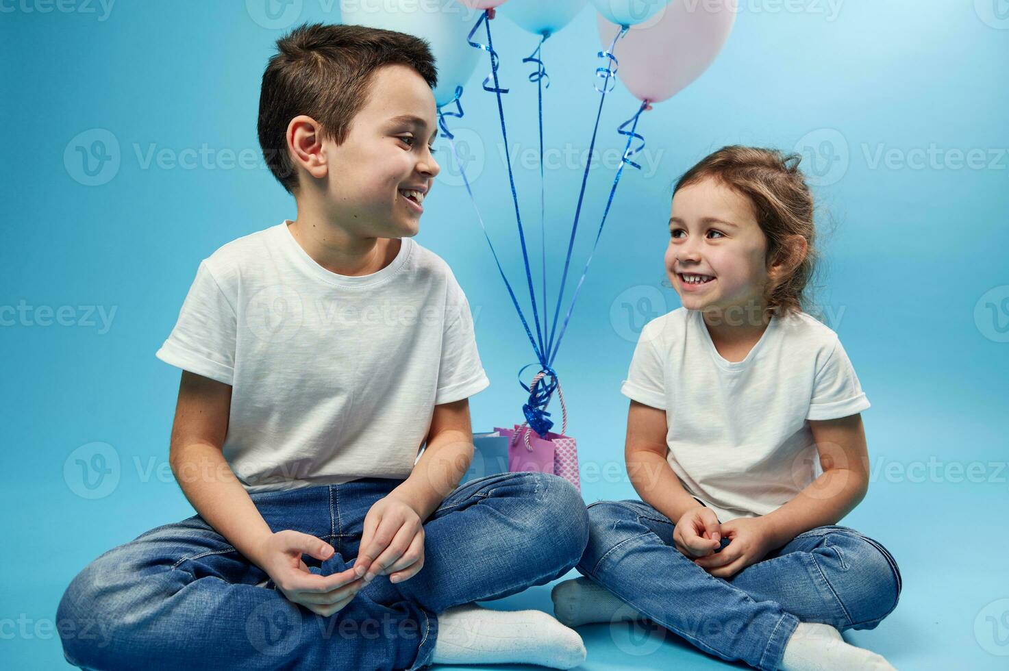 Happy children, brother and sister sit side by side and look at each other with tenderness and love on a blue background with copy space. photo