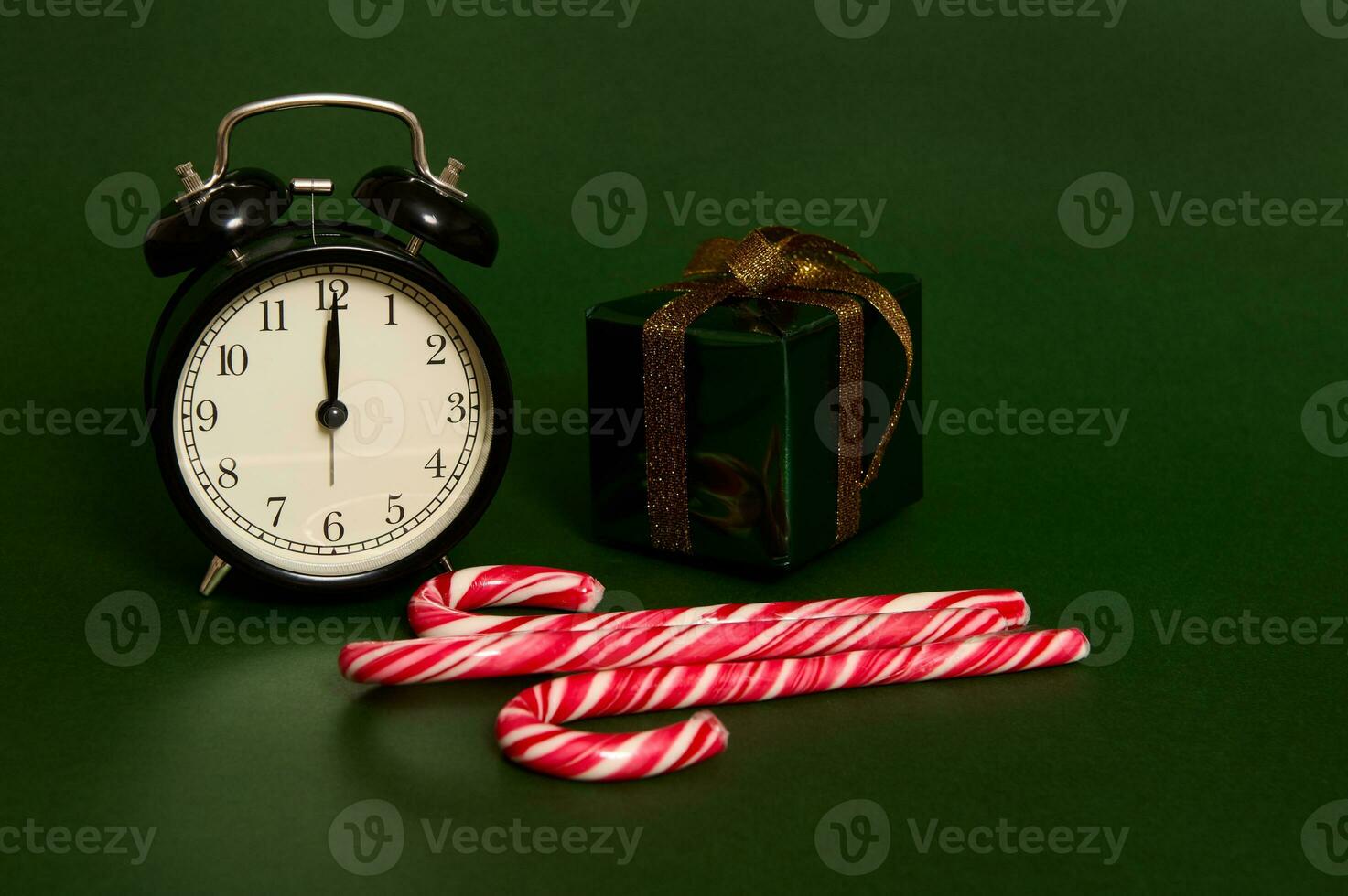 Close-up of black alarm clock with midnight on the dial, sweet striped lollipops, candy canes and Christmas gift in glitter wrapping paper and golden bow isolated on green background with space for ad photo