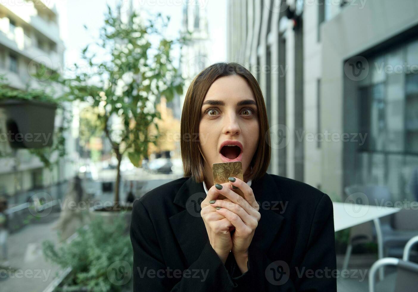 Amazed pretty woman holding gold credit card near her face, expresses astonishment, poses with opened mouth on the background of modern high-rise buildings background. Business and finance concept photo
