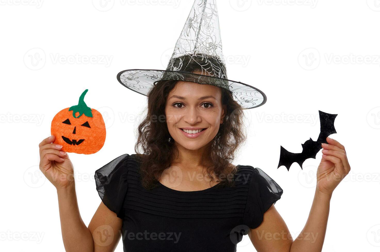 Beautiful woman in a witch hat smiles with a cute toothy smile posing against a white background with a pumpkin and bat handmade felt cutout celebrating a traditional Halloween event. Copy ad space photo