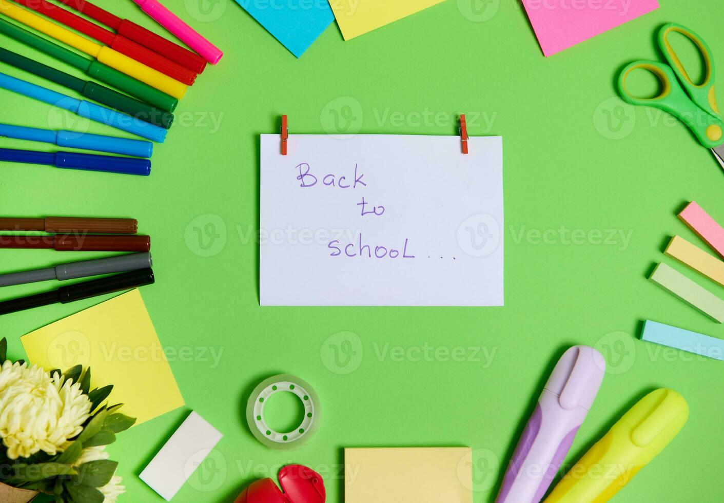 Top view of colorful assortment of stationery office supplies and school accessories arranged in a circle, with white paper in the center with lettering Back To School. Green background, copy space photo