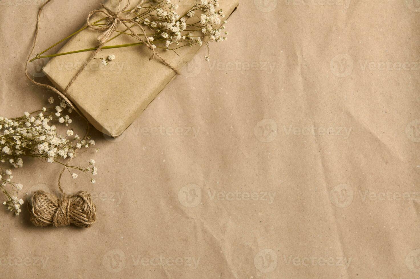 Flat lay spring composition with white gypsophila sprig on gift box in kraft wrapping paper and tied rope bow on the corner of a crumpled cardboard background with copy space for advertisement photo