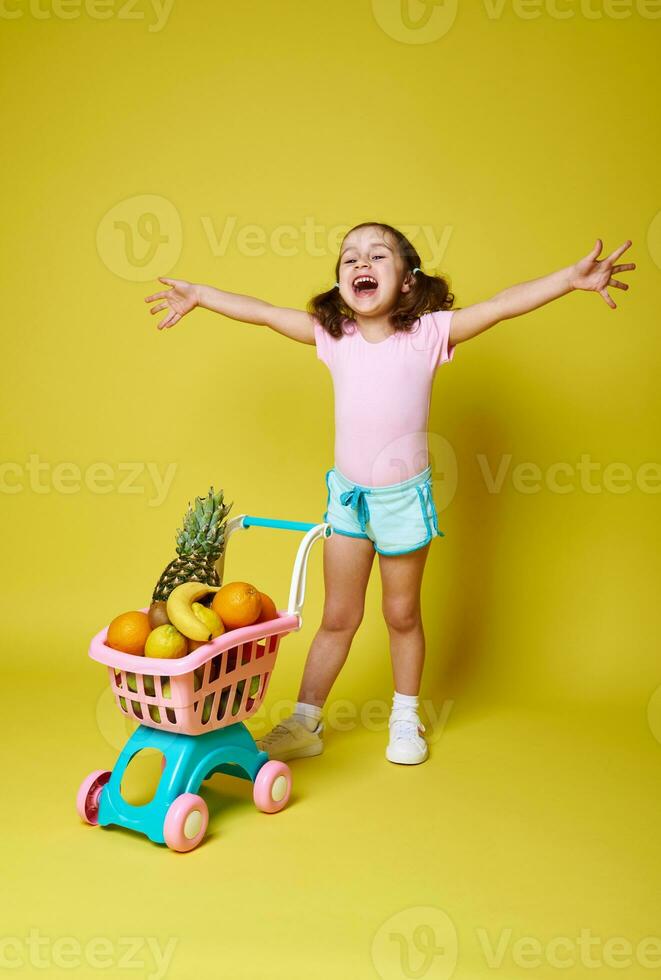 Happy little girl, laughing and holding out her hands, stands on a yellow background near her shopping trolley full of delicious exotic fruits. Emotion, happiness, childhood, healthy food concept photo