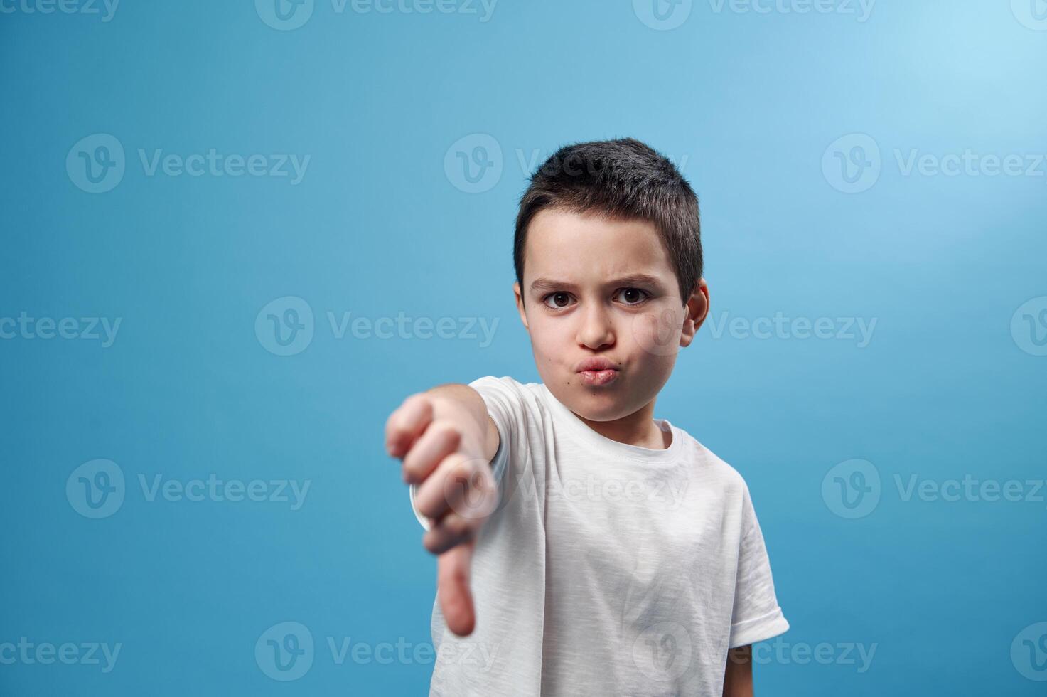 Schoolboy showing a thumb down at camera. Facial emotions on blue background with copy space photo