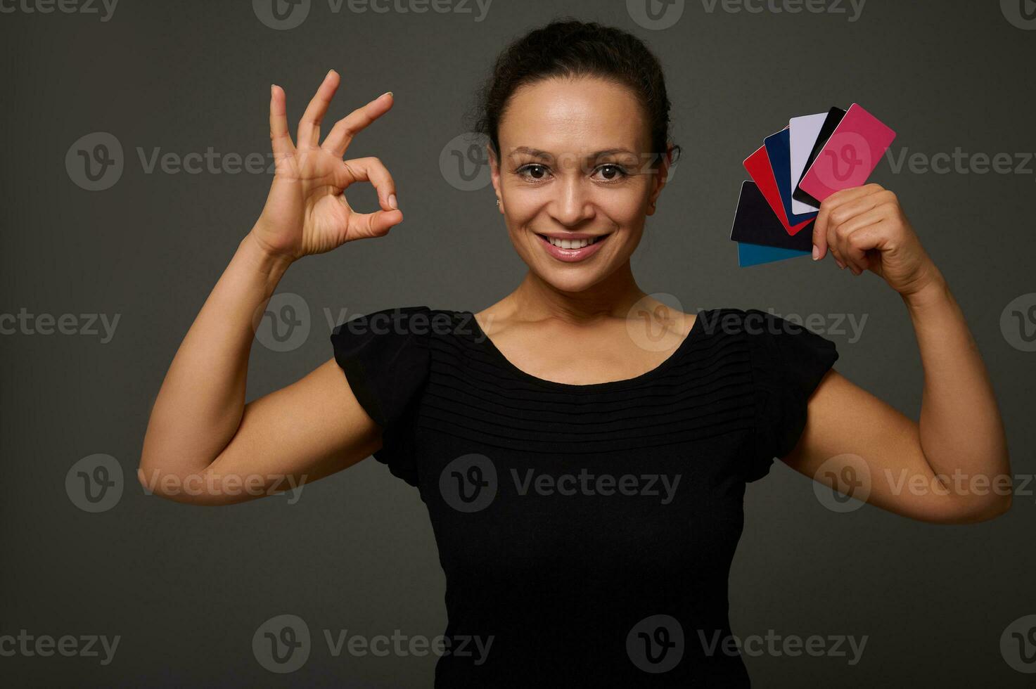 hermosa Hispano mujer sonrisas con con dientes sonrisa y poses en contra gris antecedentes con muchos descuento o crédito tarjetas en su manos demostración Okay firmar mirando a cámara. compras concepto a negro viernes foto