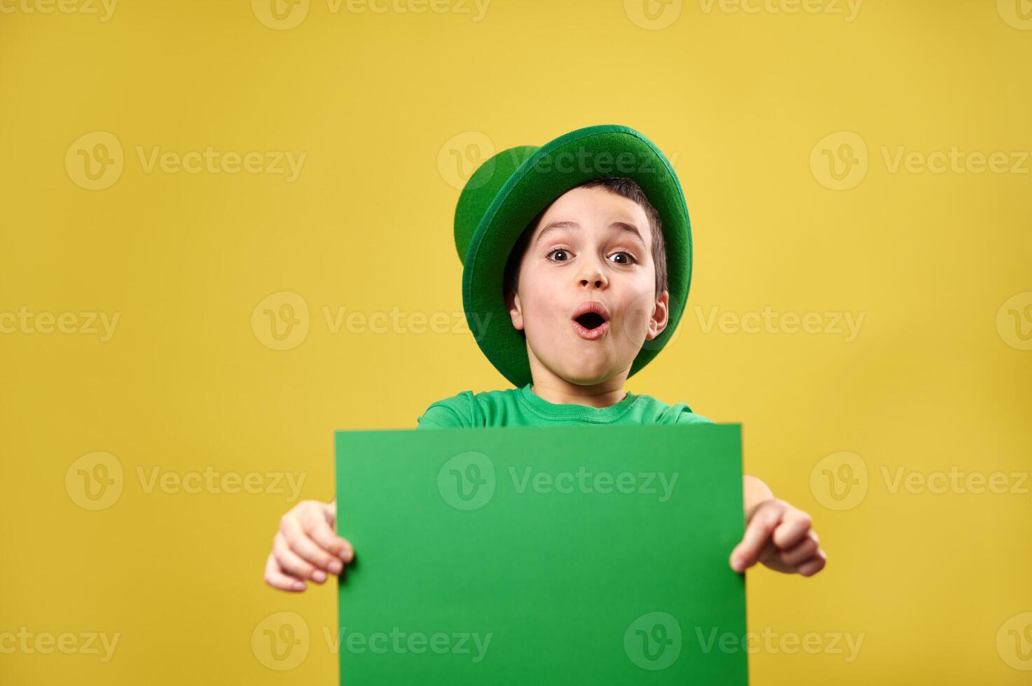 Surprised boy in green Irish leprechaun hat poses wit a green sheet of paper in his hands. Saint Patrick's Day concept. Yellow background. Copy space. photo