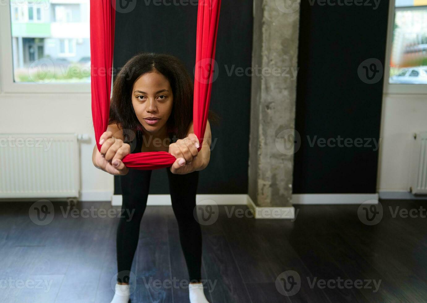 Slim body beautiful African sporty woman in black activewear stretching her arms and back muscles using red hammock during aerial flying yoga photo