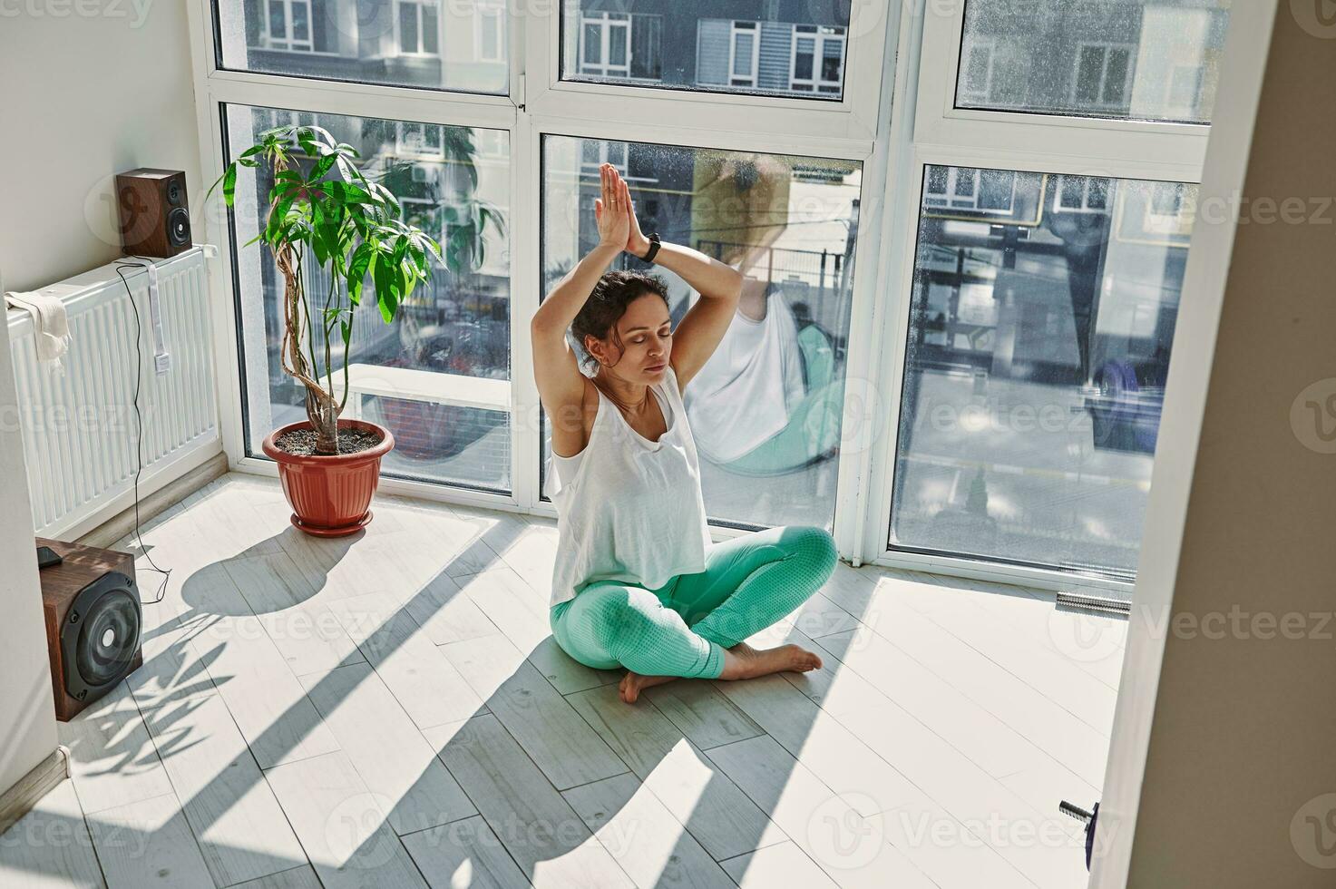 atento joven mujer en ropa de deporte sentado en padmasana loto posición con elevado brazos disfrutando cerrado ojos meditación durante yoga práctica a hogar foto