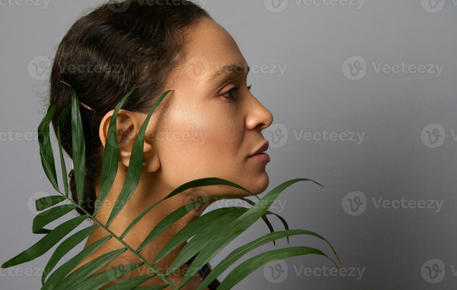 Close-up side portrait of face of beautiful woman with healthy clean bright skin posing with palm leaf on gray background. Copy ad space. Cosmetology beauty skin care anti-aging treatment rejuvenation photo