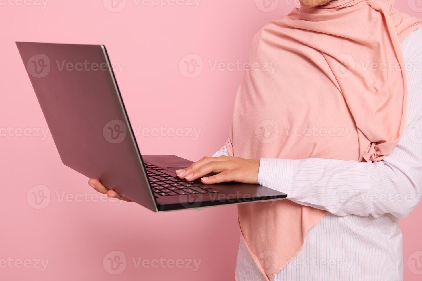 Close-up portrait of a woman wearing pink hijab standing against colored background with an open laptop in hands and typing text.Concept of successful Muslim programmer woman, worker at IT technology photo