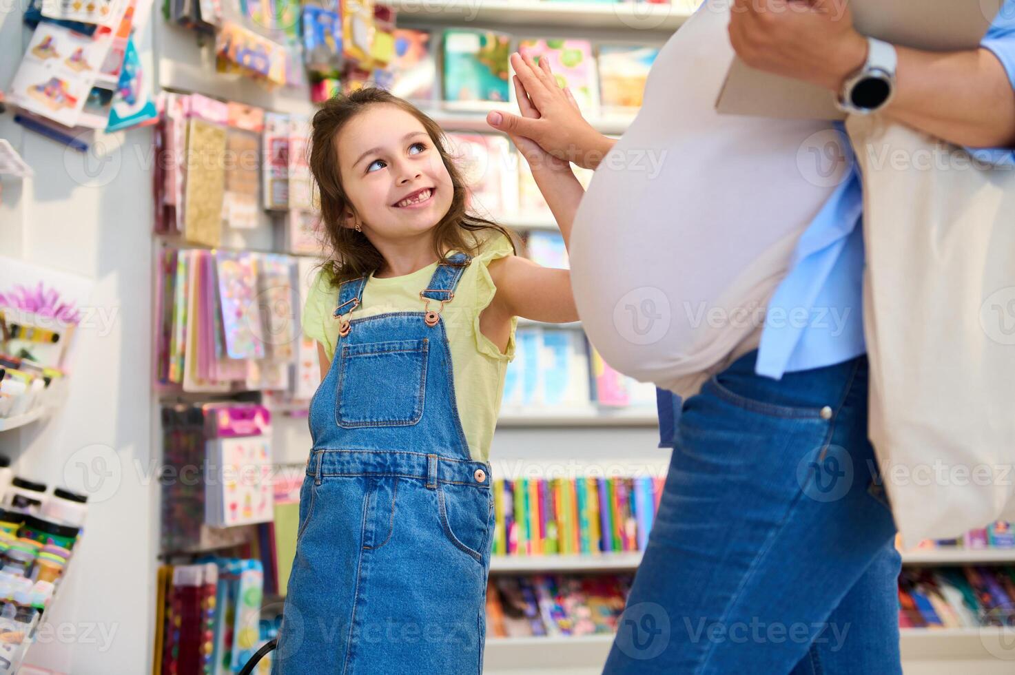 Cheery smiling kid girl gives high five to her mother while shopping together for school supplies in stationery store photo