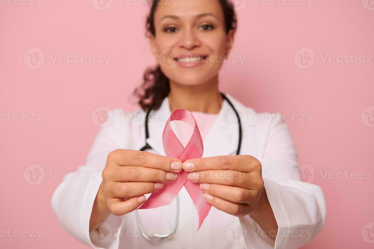 Soft focus on Breast Cancer Awareness Pink Ribbon, in hands of blurred mixed race female doctor in white medical coat, isolated on colored background. 1 st October, World Day of fight breast cancer photo