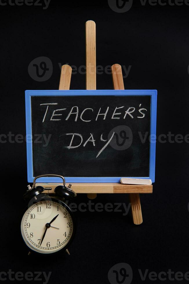 Close-up of a chalk board with lettering Teacher's Day, standing on a wooden table easel, next to black alarm clock, isolated on black background with copy space. photo