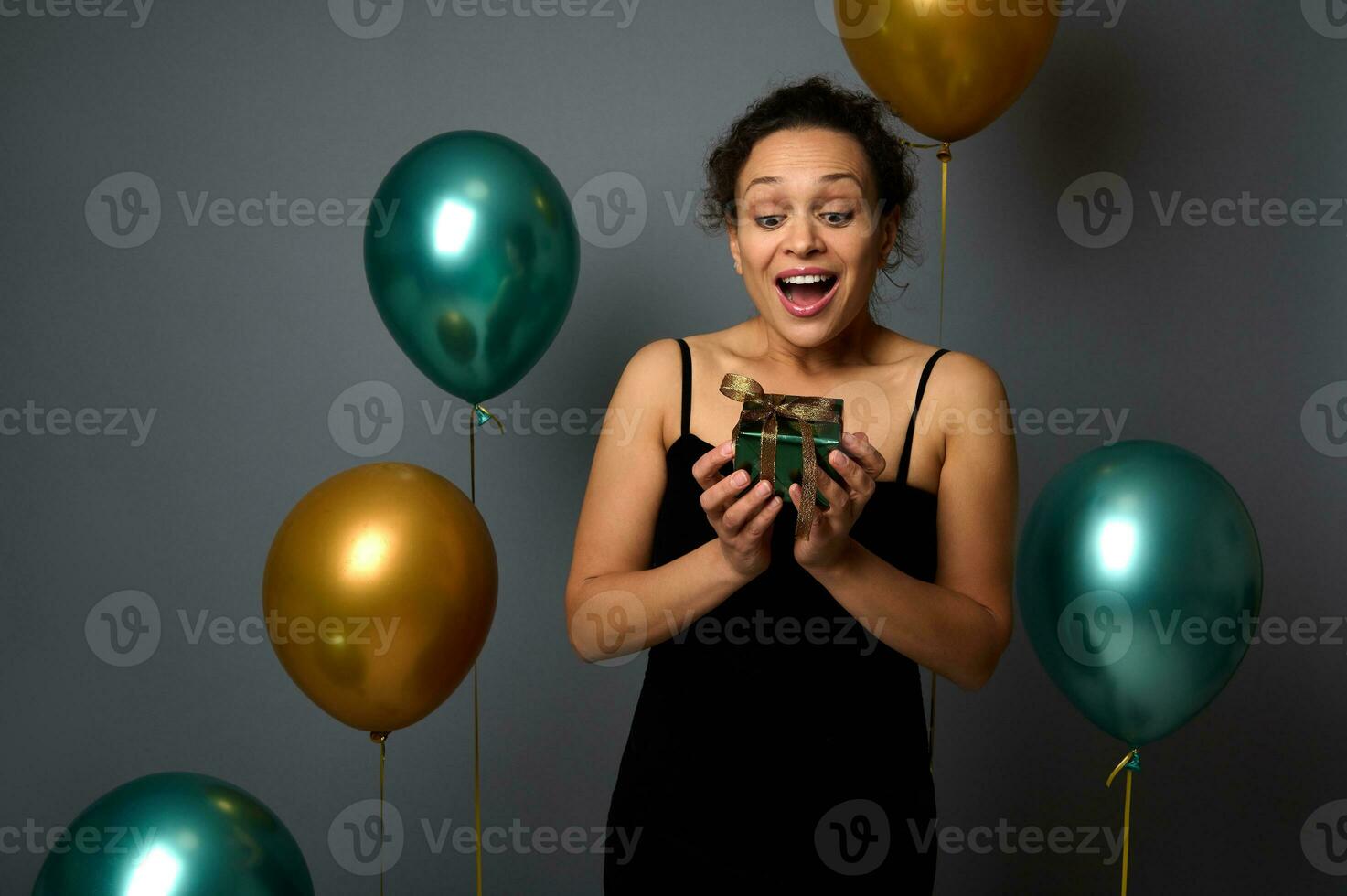 Gorgeous African woman wearing an evening black dress, surprised by a Christmas gift in shiny green wrapping paper and golden bow, isolated over gray background with gold and green metallic air balls photo